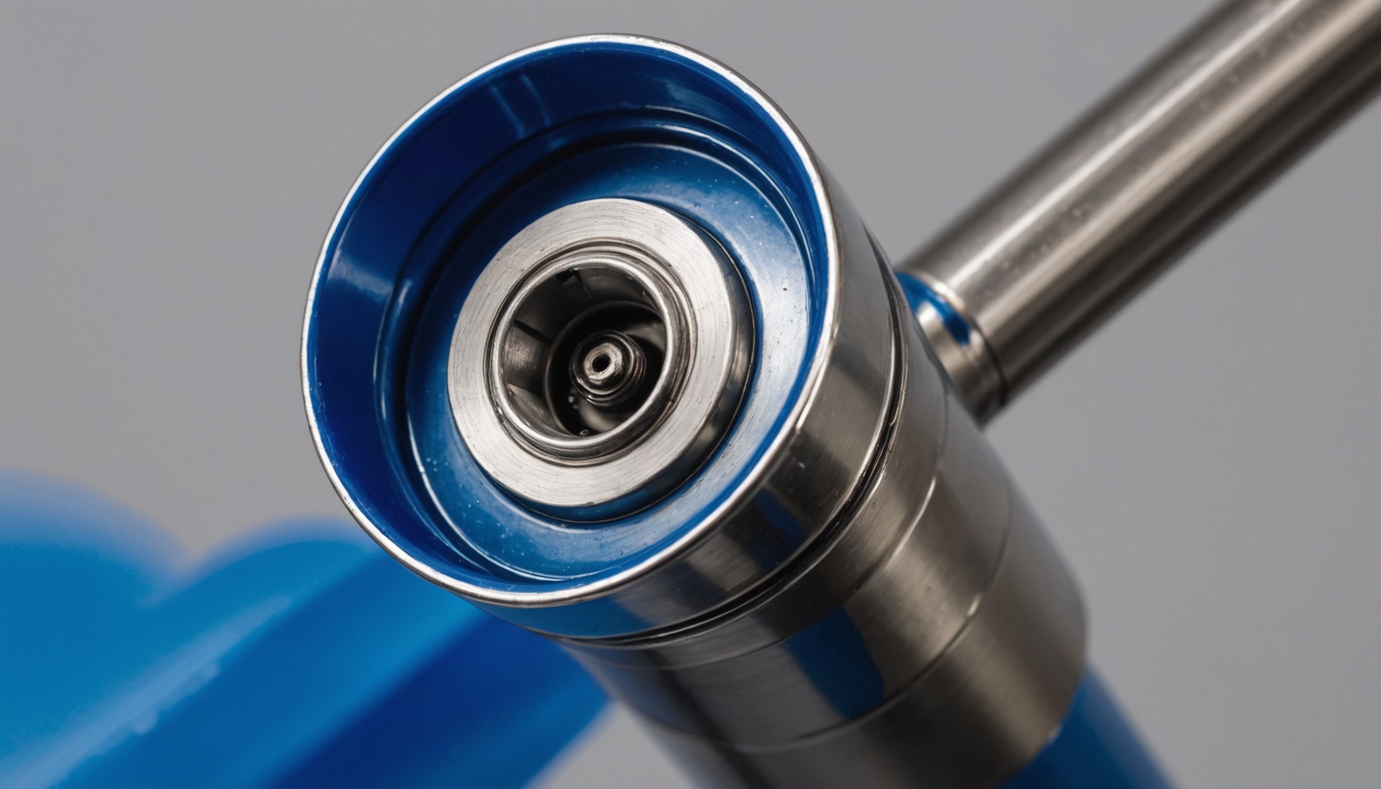 An extreme close-up of the air cap and nozzle of an automotive spray gun, the polished steel cap filling the centre of the frame with the fluid tip at its centre, a band of deep blue paint drawn across the cap, and the blue body of the gun running away below it against a plain grey background. Both of the airflow requirements a spray booth answers to arise at this opening. The paint leaving it becomes the overspray that a velocity across the working section has to carry away from the operator, and the solvent evaporating out of it becomes the vapour that a separate airflow has to dilute below a fraction of its explosive limit. The first requirement follows from the size of the booth and the second from what comes through this nozzle and how fast, so neither of the two can be read off the other