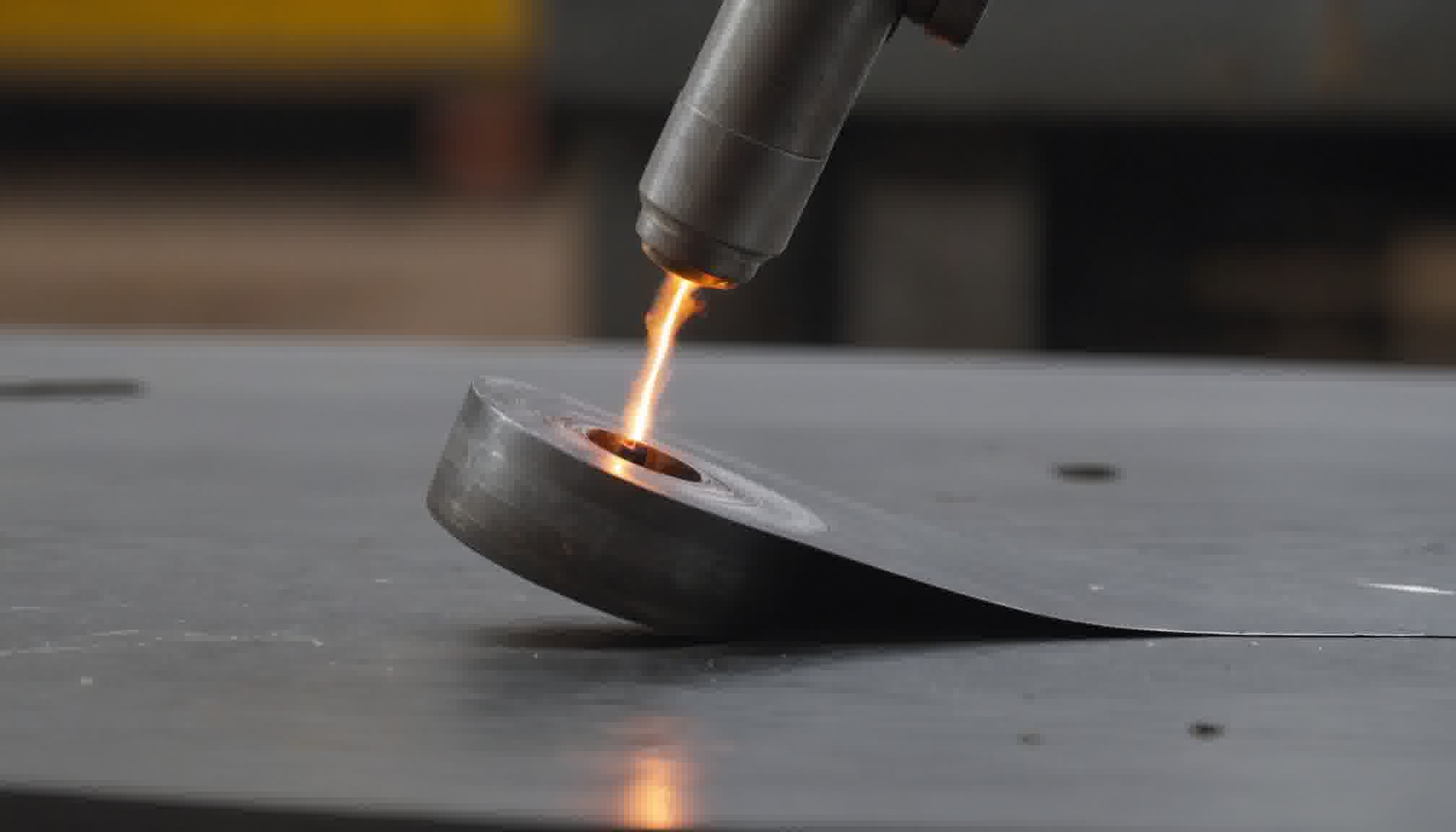 An extreme close-up of a cutting torch working on a steel plate in a dark workshop, the barrel of the torch entering from the top of the frame and a narrow jet of intense orange flame running from its tip down to a small molten pool, the plate lifting and curling away from the cut with the heat, the background reduced to soft dark shapes. The glowing point is the whole of the problem an extraction calculation is trying to solve: fume is created here, in a spot a few millimetres across, and it starts rising immediately. What decides whether a hood collects it is not the size of the hood or the airflow through it but the distance between this point and the hood face, which enters the requirement squared and appears nowhere in a face-area calculation